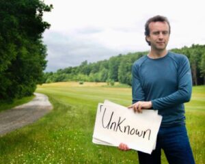 Eric Everett on dirt country road holding a sign with the word Unknown