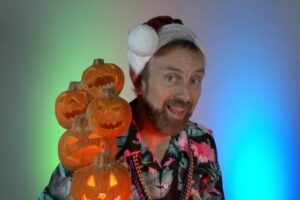 Eric Everett holding a stick of stacked pumpkins, wearing Santa hat and colorful Hawaiian shirt